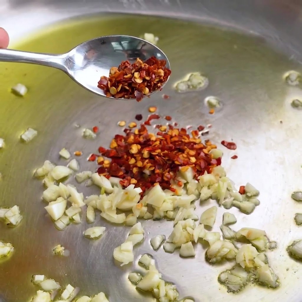 Gehackter Knoblauch und rote Chiliflocken brutzeln in einer Edelstahlpfanne mit Olivenöl.