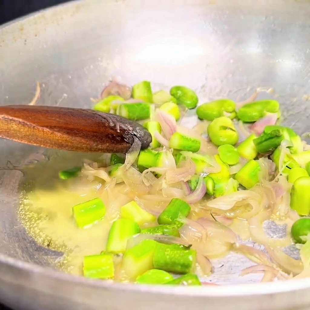 Angedünstete Zwiebeln und grünes Gemüse werden mit einem Löffel aus Metall aus einer Metallpfanne geschöpft.