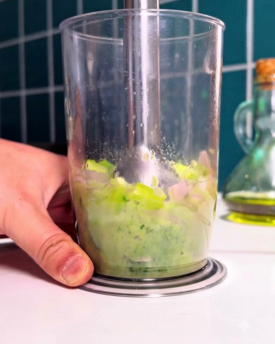 Ein Stabmixer püriert blanchiertes grünes Gemüse und Spinat in einem hohen durchsichtigen Becher.