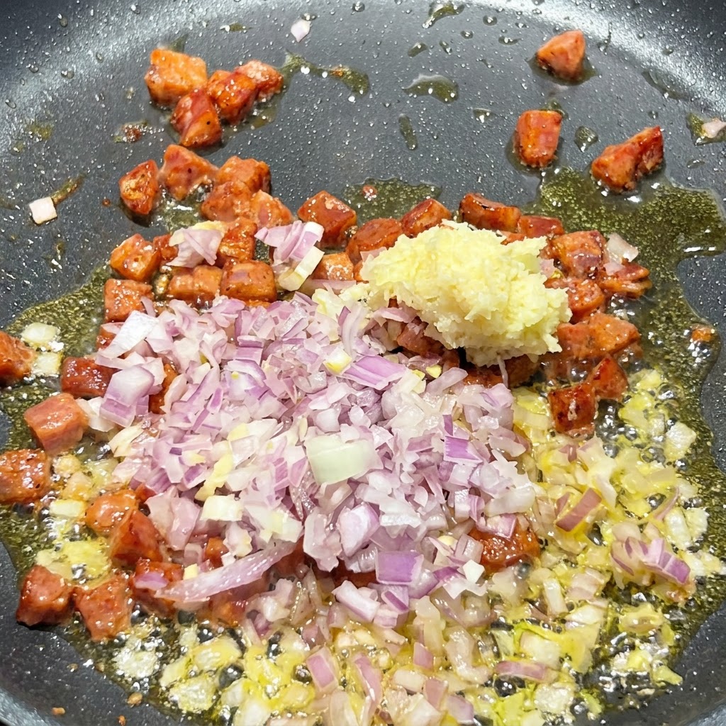 Gewürfelte rote Zwiebel und gehackter Knoblauch werden in die brutzelnde Pfanne mit angebratenen Chorizostücken gegeben.