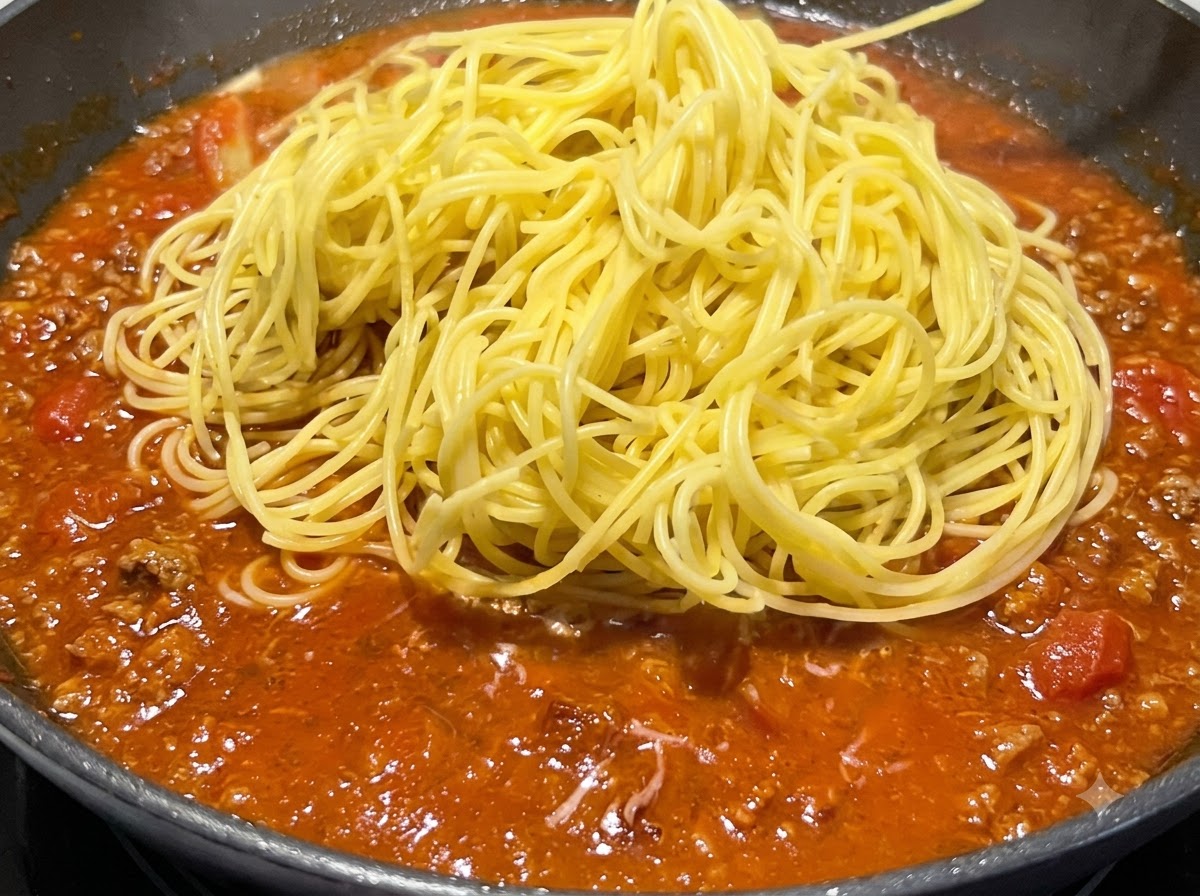 Ein Haufen gekochter gelber Spaghetti liegt auf der köchelnden roten Soße in einer Pfanne.