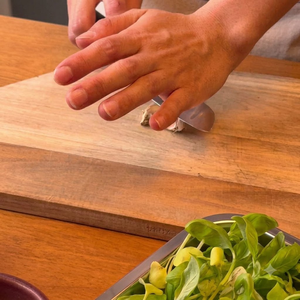 Eine Hand drückt die flache Seite eines Kochmessers auf eine Knoblauchzehe auf einem Holzschneidebrett.