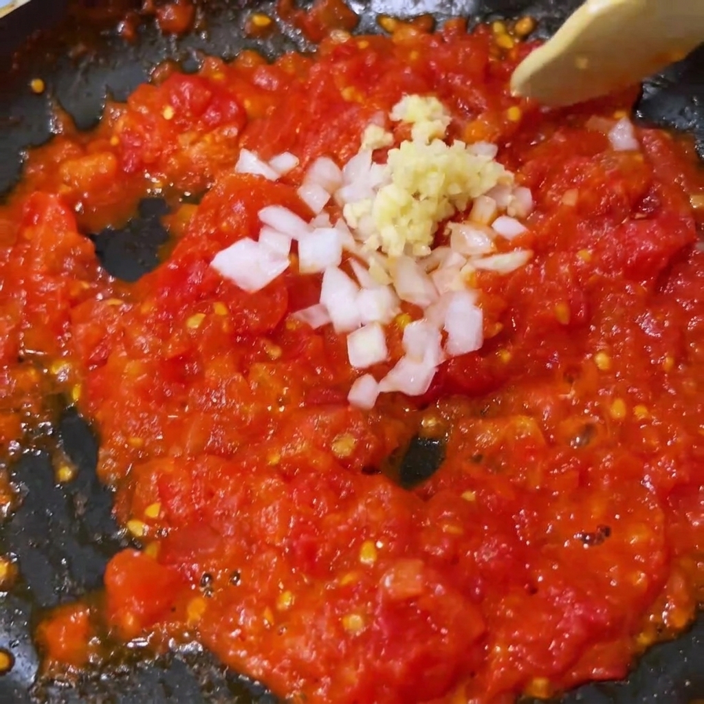 Gehackter Knoblauch und weiße Zwiebel werden in einer Pfanne unter weichgekochte Tomaten gemischt.