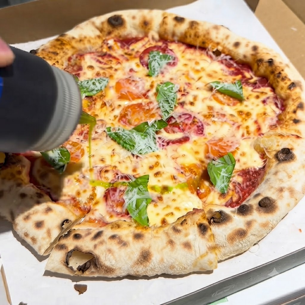 Eine Flasche gießt einen Strahl natives Olivenöl extra auf eine frisch gebackene Pizza, die im Karton mit Basilikum und Käse belegt ist.