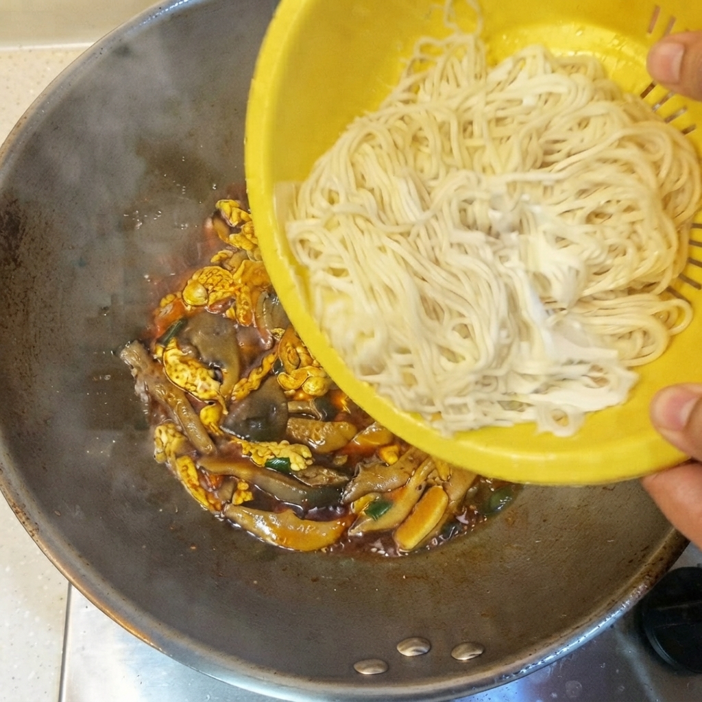 Gekochte dünne Nudeln werden aus einem gelben Sieb in den Wok mit Sauce und Zutaten gefüllt.
