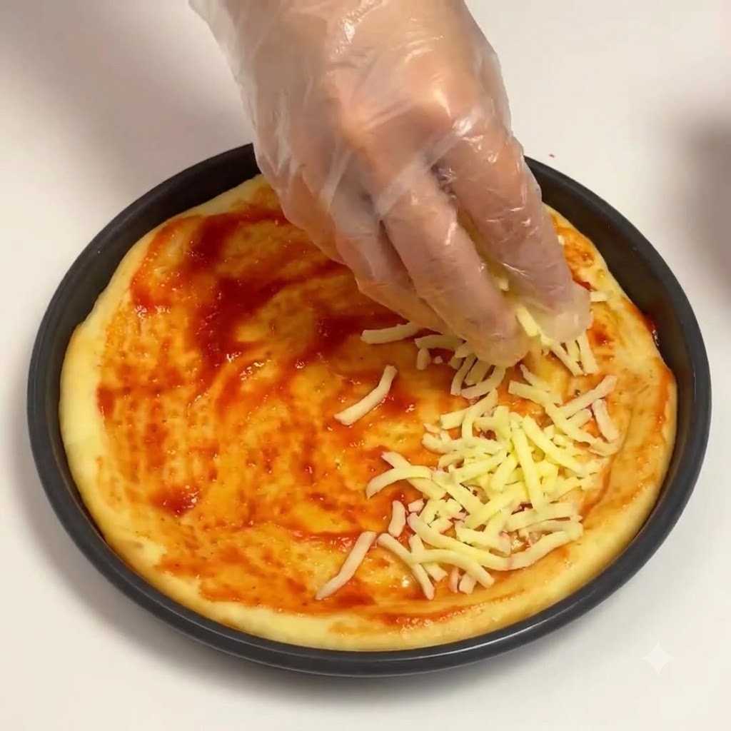 Eine behandschuhte Hand streut geriebenen Mozzarella-Käse über die Tomatensoßenbasis in einer runden Pizzapanne.