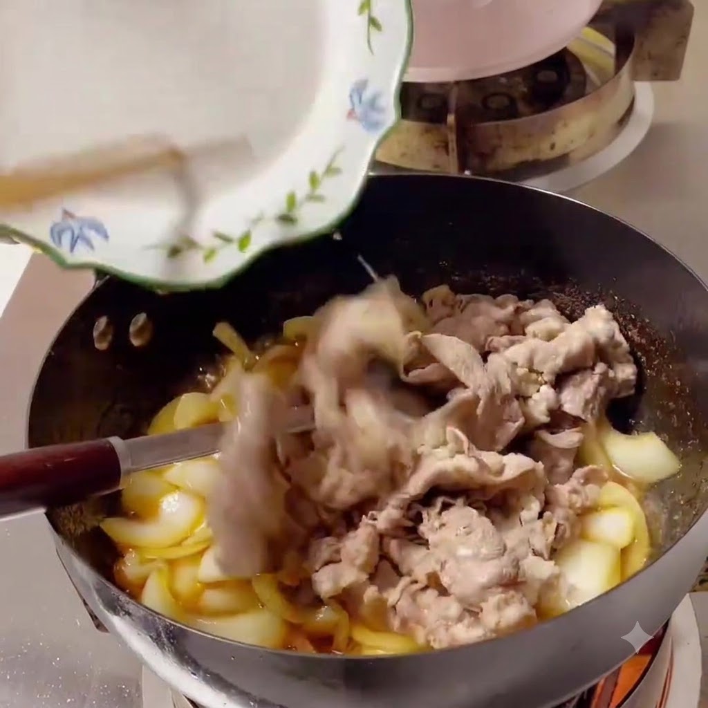 Gekochte Rindfleischscheiben werden von einem weißen Teller in einen mit Zwiebeln und Sauce gefüllten Wok geschoben.