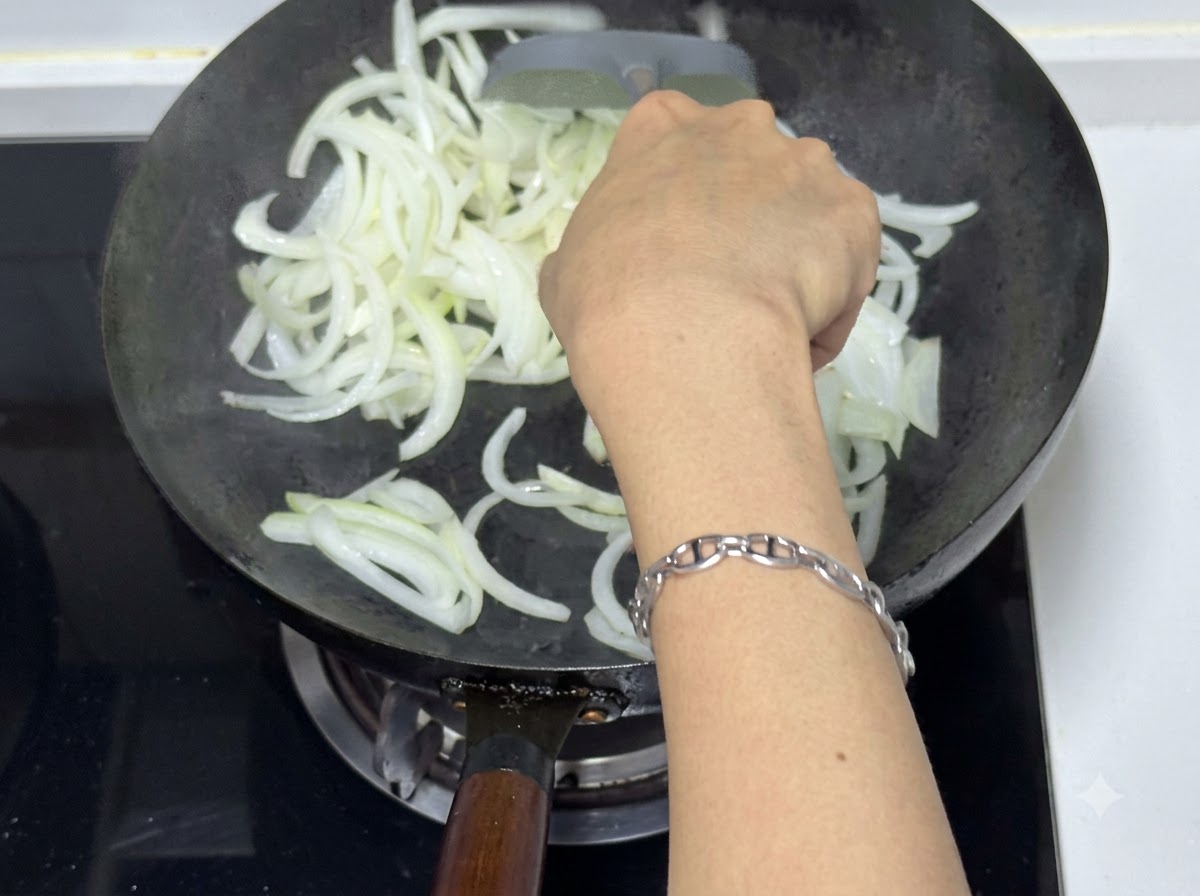 Eine Person benutzt einen Pfannenwender, um geschnittene Zwiebeln in einem Wok anzubraten, was durchscheinende und leicht gebräunte Stücke zeigt.