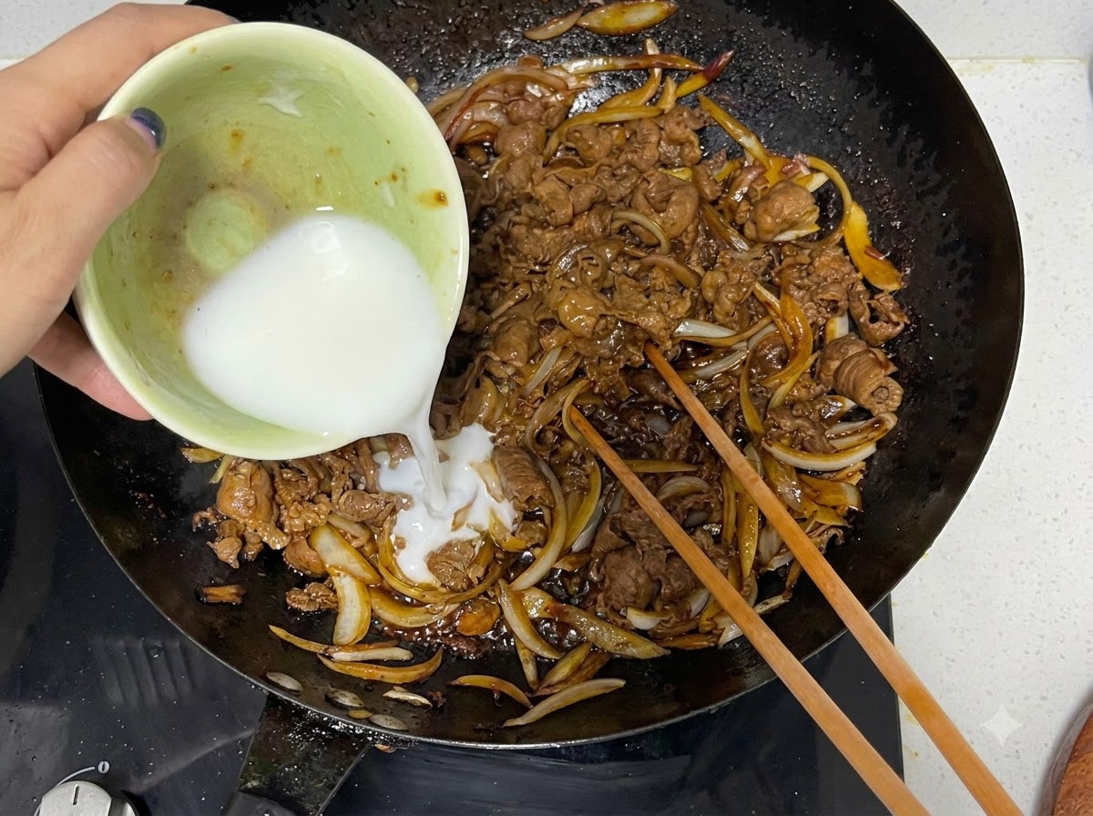 Maisstärkemischung wird aus einer Schüssel in einen Wok mit gekochtem Rindfleisch und Zwiebeln gegossen, daneben liegen Essstäbchen aus Holz.