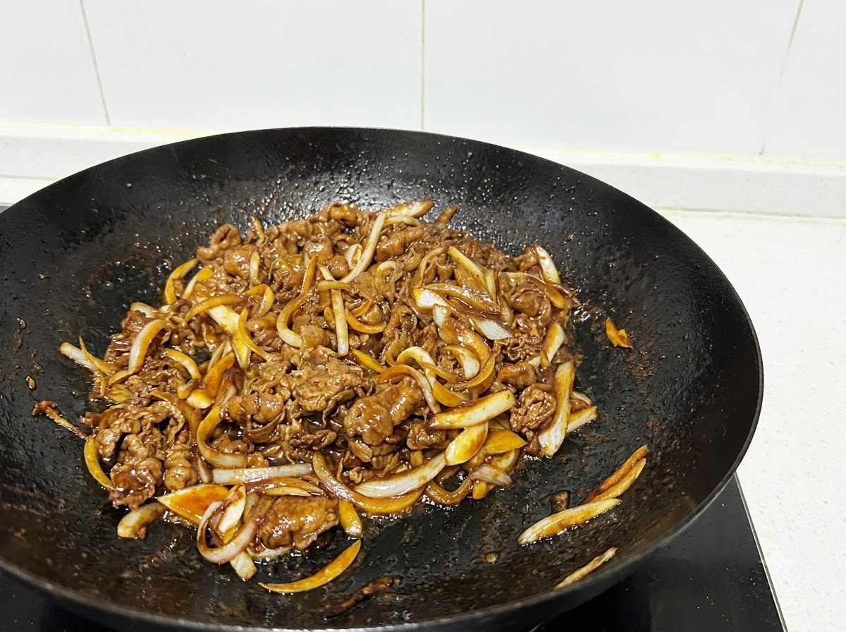 Eine Nahaufnahme von Rindfleischstreifen und Zwiebelstreifen, die in einem schwarzen Wok mit Kochutensilien angebraten werden.