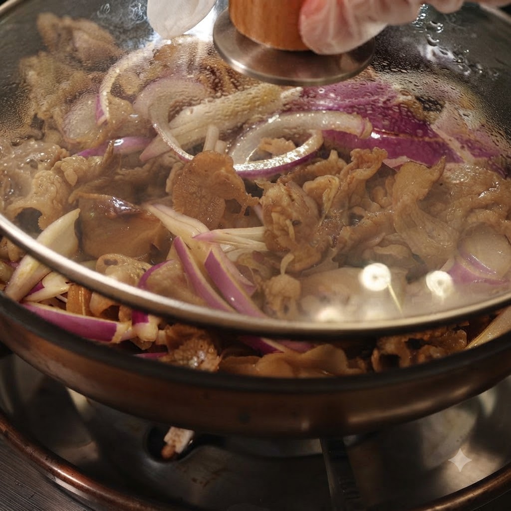 Eine Pfanne, abgedeckt mit einem klaren Glasdeckel, zeigt köchelndes Rindfleisch und rote Zwiebeln im Inneren.