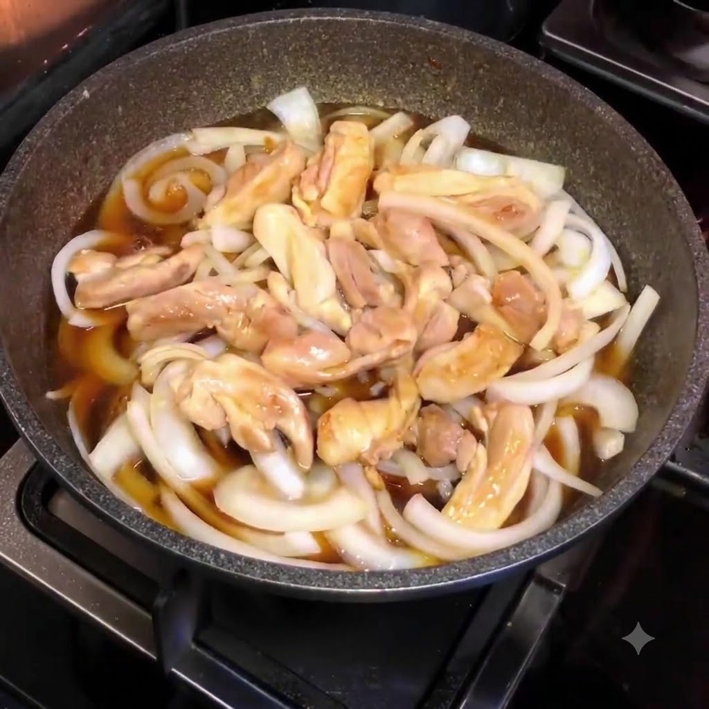 Eine Pfanne gefüllt mit Zwiebeln, Hähnchenstücken und brauner Soße, von oben während des Kochvorgangs fotografiert.
