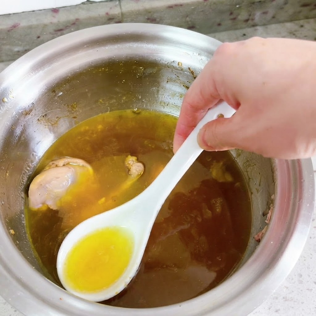 Ein weißer Löffel schöpft hellgelbes Öl von der Oberfläche einer köchelnden Entensuppe ab.
