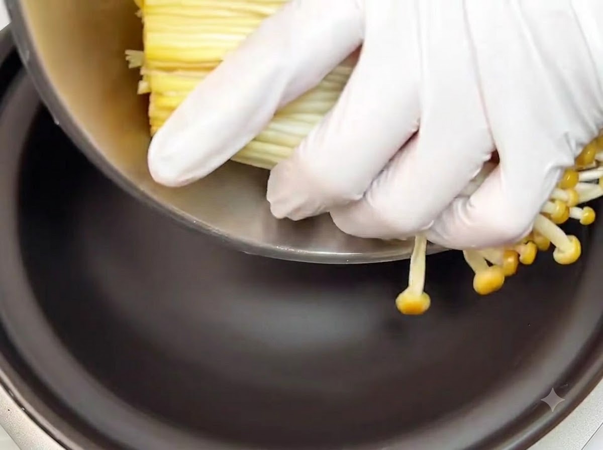 Mit Handschuhen versehene Hände legen ein Bündel frisch geschnittener Enoki-Pilze auf den Boden eines schwarzen Tontopfs.