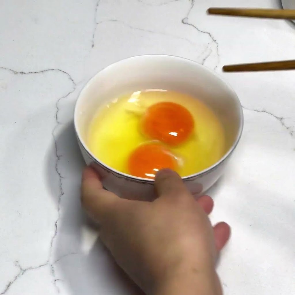 Zwei frische Eier in einer weißen Schüssel, die mit Holzstäbchen ohne Gewürze verquirlt werden.