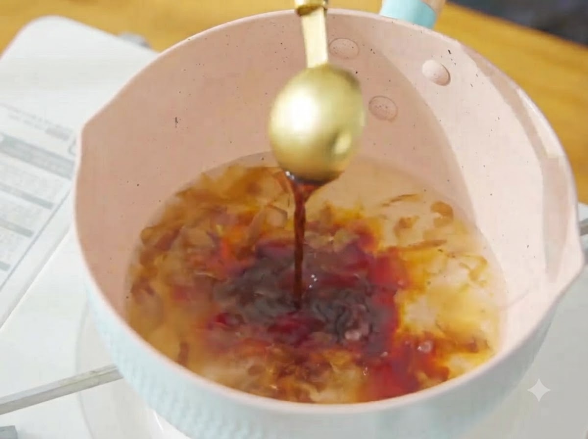 Dunkle Sojasauce wird von einem goldenen Löffel in die köchelnde Bonito-Brühe im rosa Topf gegossen.