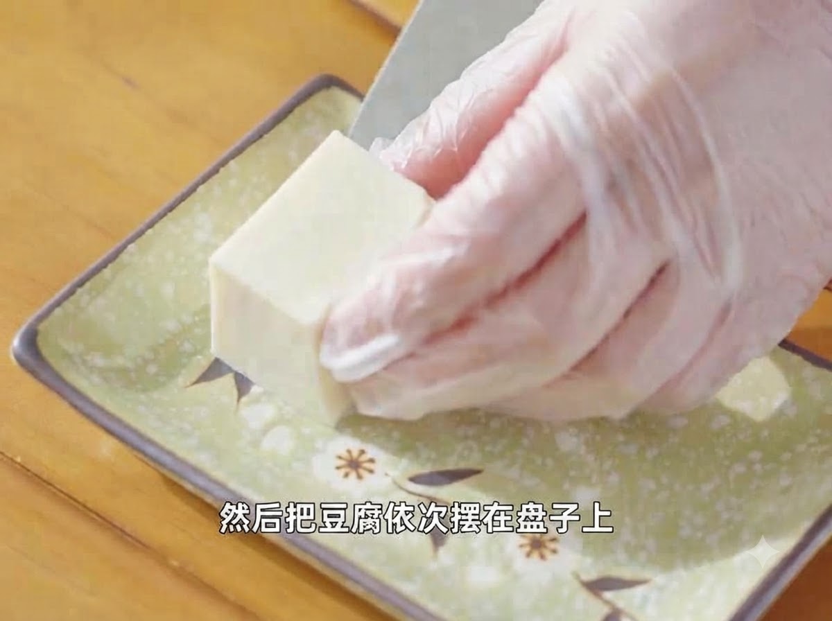 Behandschuhte Hände ordnen ein Stück geschnittenen Seidentofu auf einem gemusterten grünen Servierteller an.