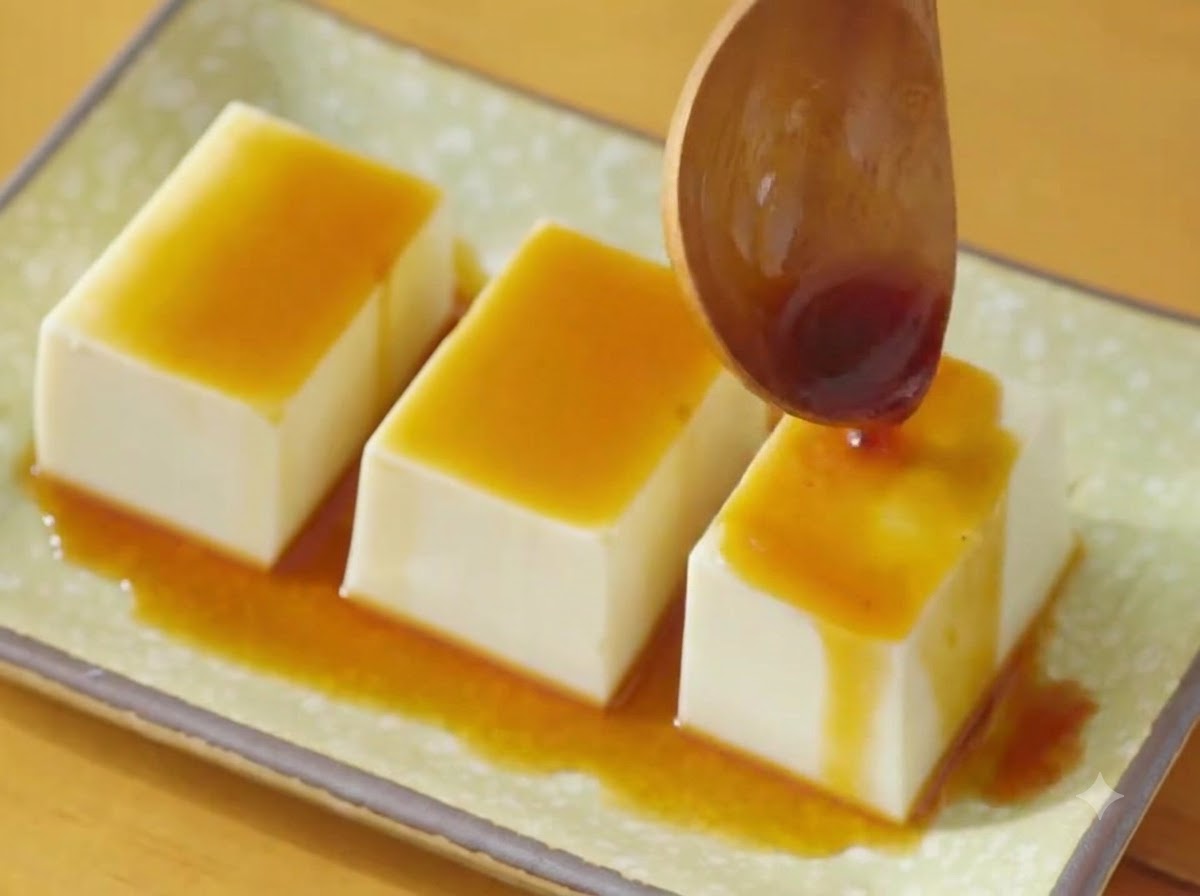 Ein Holzlöffel gießt dunkle Sojasauce über drei Reihen Seidentofu auf einem grünen Teller.
