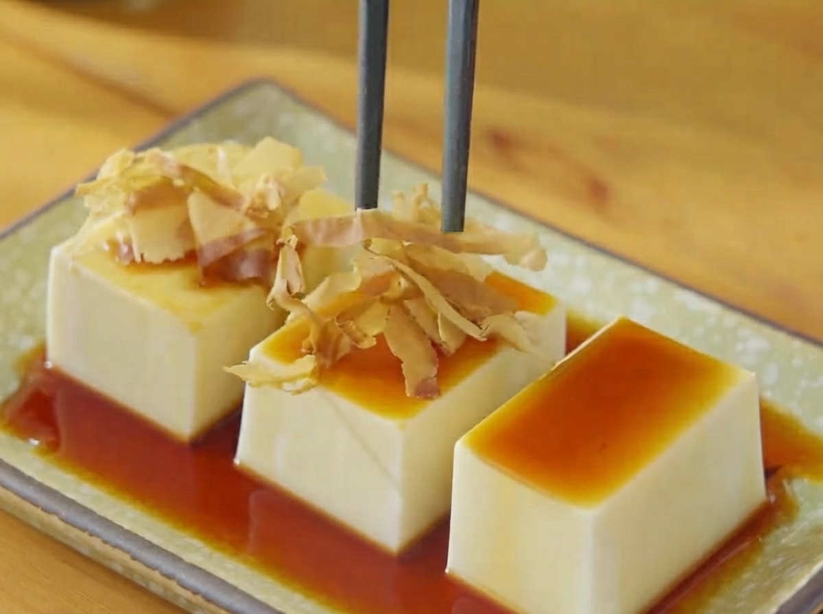 Essstäbchen platzieren trockene Bonitoflocken und Algenstreifen auf dem mit Sojasauce beträufelten Tofu.