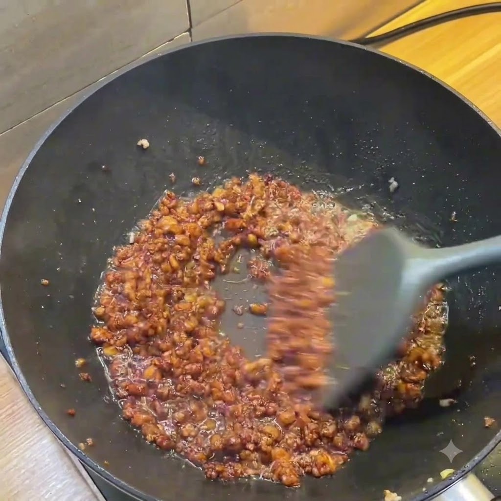 Ein dunkler Wok mit einer reichhaltigen, braunen Hackfleischsauce, die mit einem grauen Spatel gerührt wird.