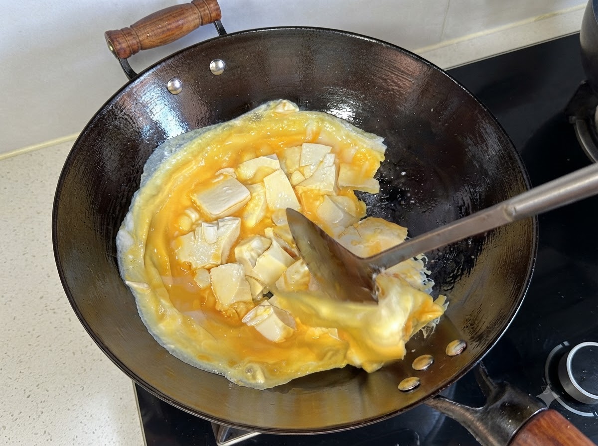 Ein Pfannenwender hebt und wendet die gegarte Schicht aus Eiern und Tofu im Wok.