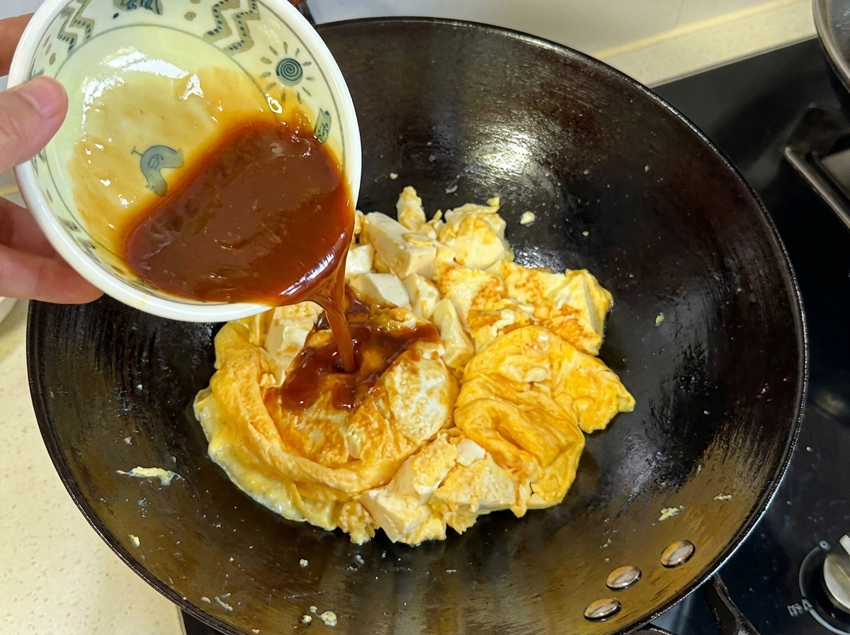 Dunkelbraune Soße wird aus einer kleinen Schüssel auf die gegarte Ei-Tofu-Mischung im Wok gegossen.