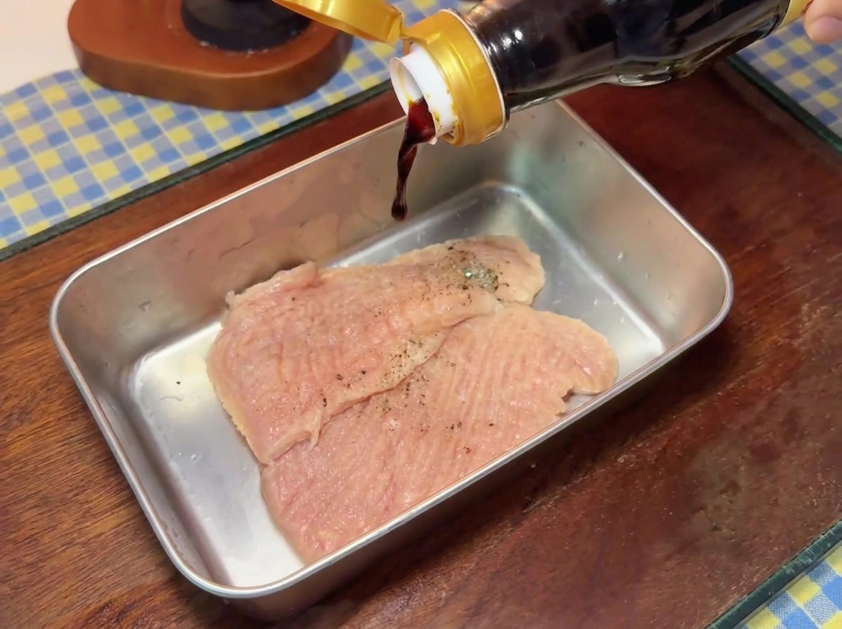 Dunkle Sojasauce fliesst aus einer Glasflasche auf gewürzte Schweineschnitzel in einer Metallschale.