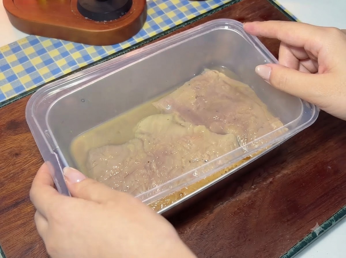 Ein Plastikdeckel wird auf einen Behälter mit marinierten Schweineschnitzeln und Flüssigkeit gesetzt.