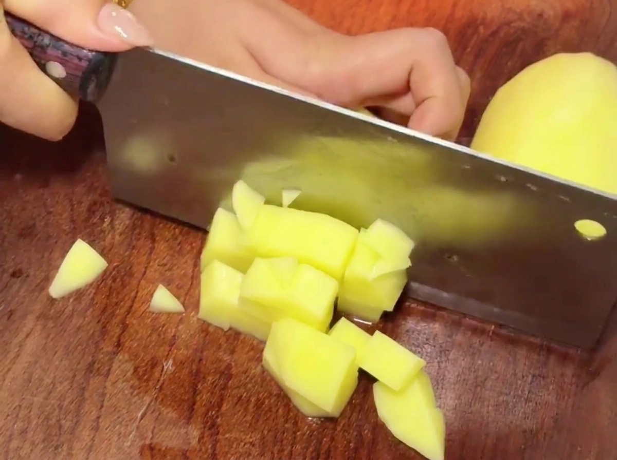 Ein Fleischbeil schneidet gelbe Kartoffelstücke auf einem Holzschneidebrett in kleine Würfel.