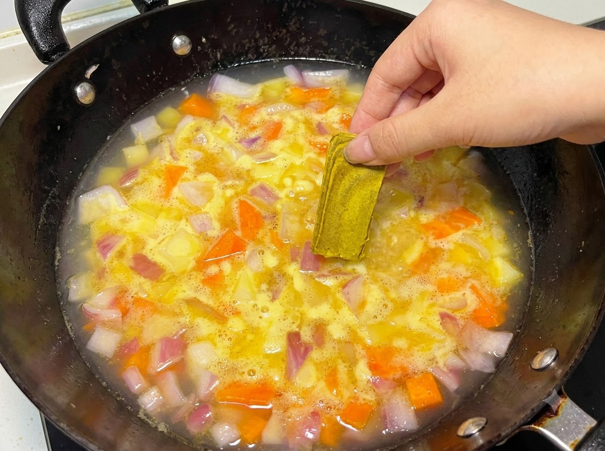 Eine Hand legt einen festen Curry-Block in einen köchelnden Topf mit Wasser und Gemüse.
