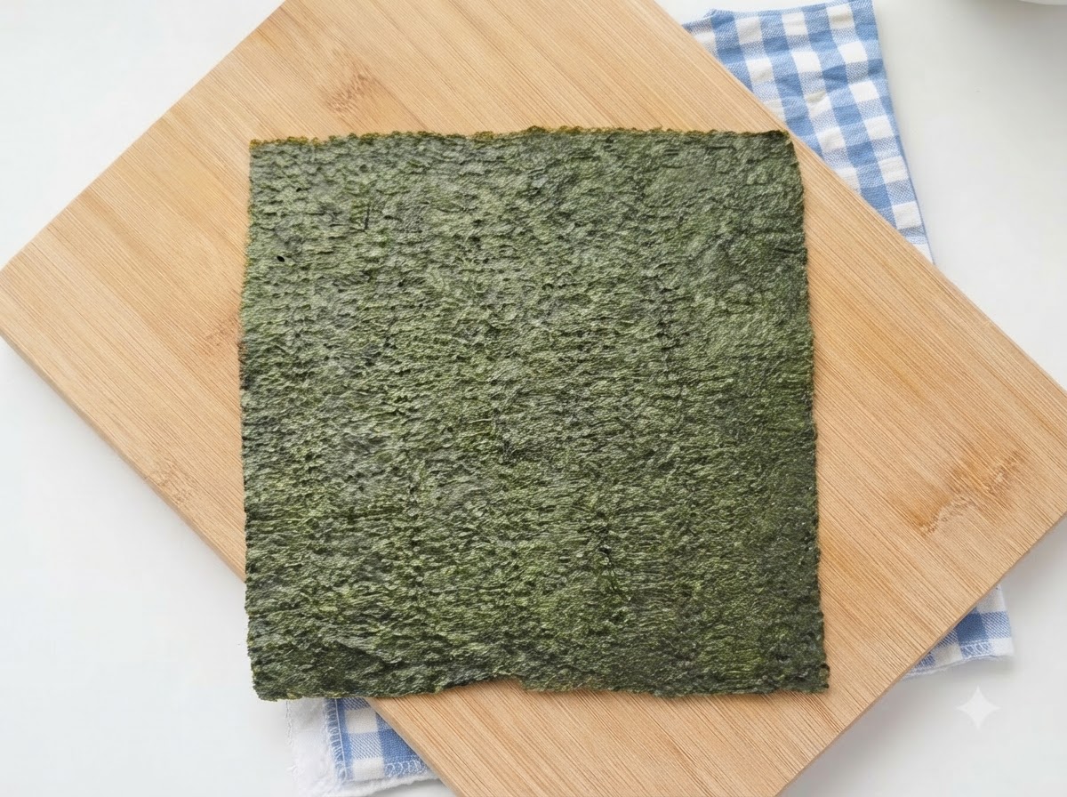 Ein vollständiges Blatt grünes Sushi-Nori liegt auf einem Holzschneidebrett mit einem blau karierten Tuch darunter.