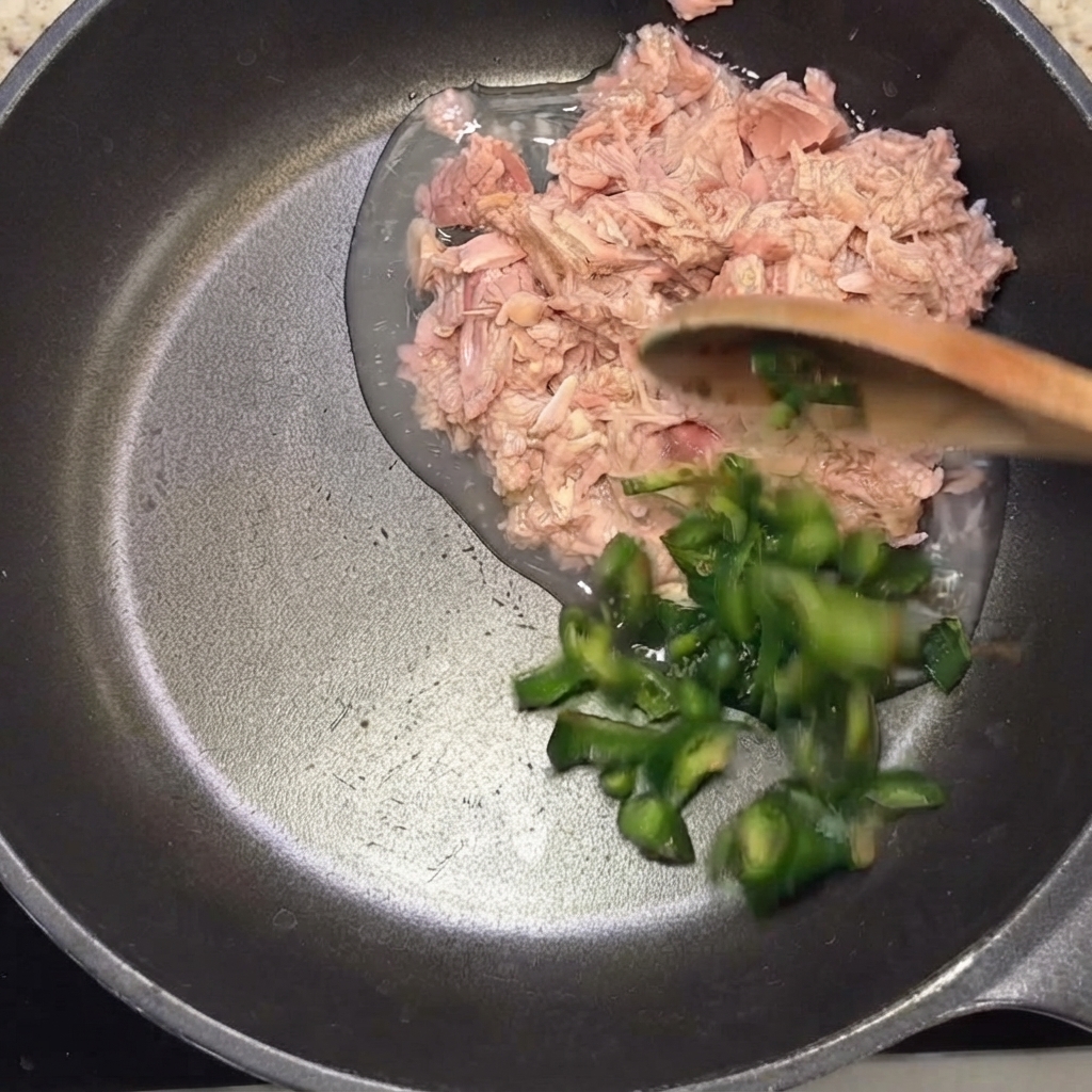 Geschnittene grüne Chilis werden mit einem Holzlöffel zum Thunfisch in die Pfanne gegeben.
