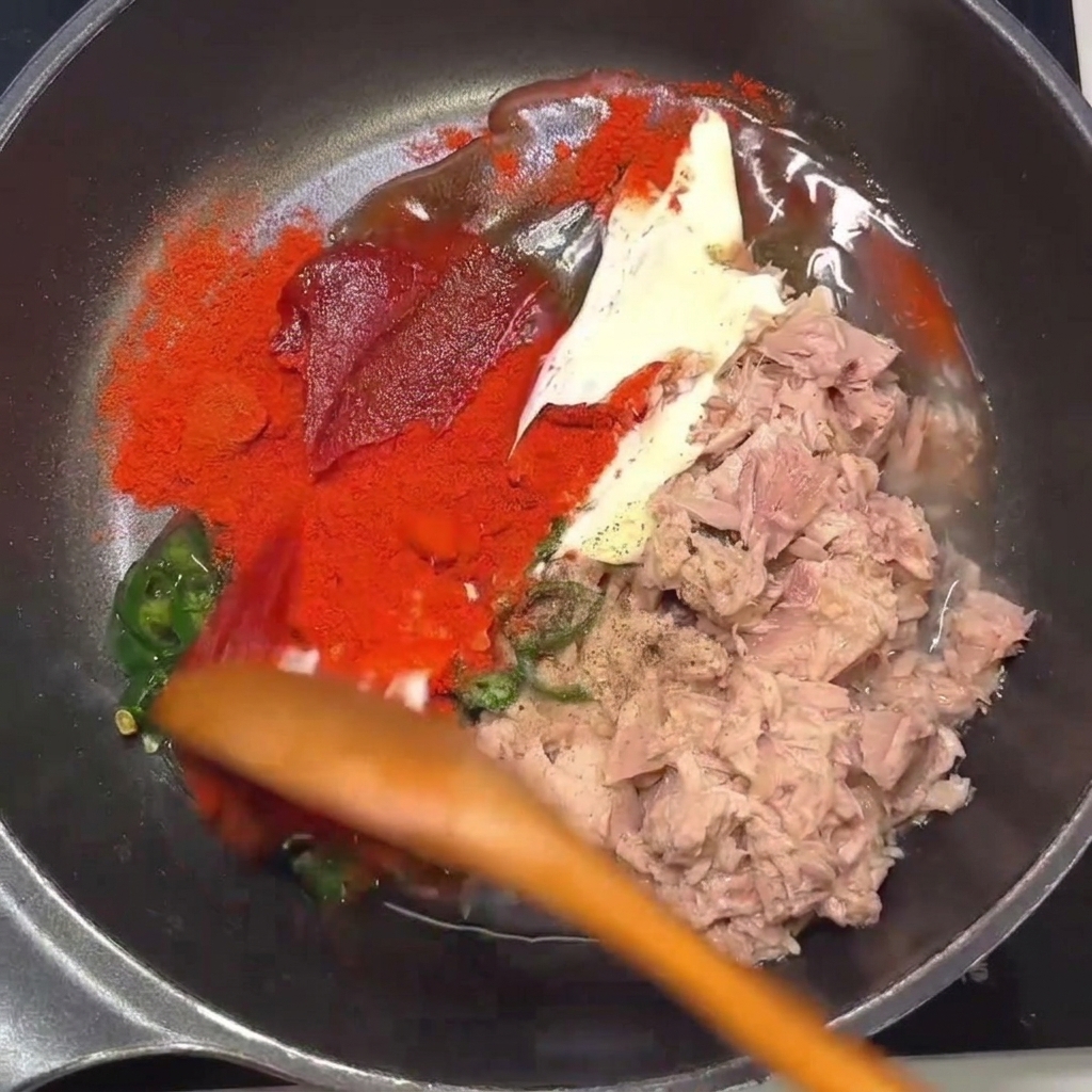 Ein Holzlöffel vermengt Thunfisch, Chili und Gewürze in einer Pfanne.