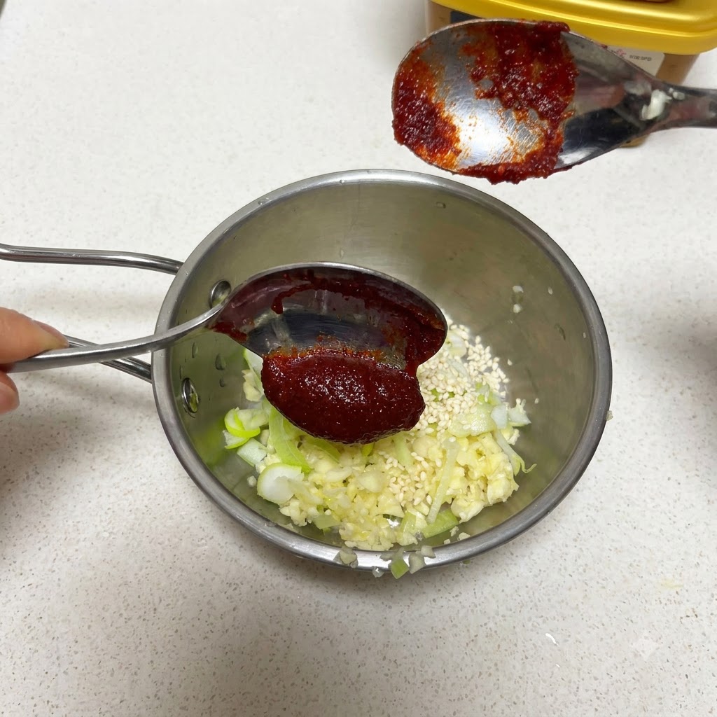 Ein Löffel koreanischer Chilipaste wird über eine Pfanne mit Knoblauch und Sesamsamen gehalten.