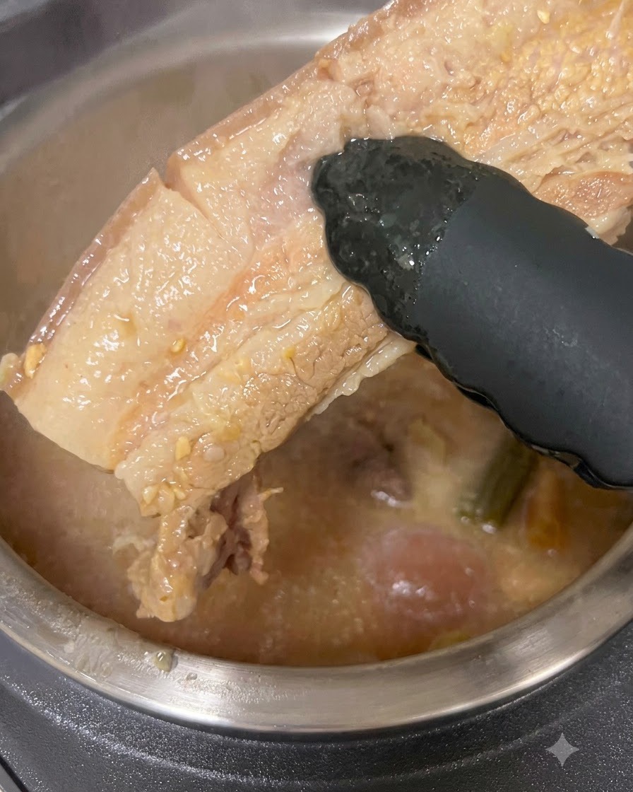 Ein dickes Stück gekochter Schweinebauch wird mit einer schwarzen Zange aus einem Topf mit Brühe gehoben.