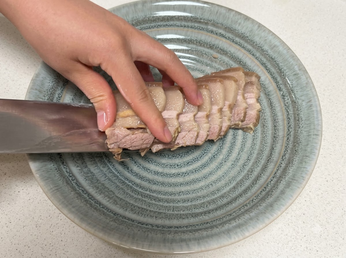Scheiben von gekochtem Schweinebauch werden mit einem Messer auf einen strukturierten grauen Teller gelegt.