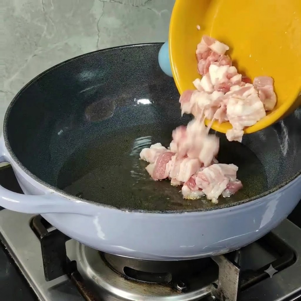 Gewürfelte rohe Schweinebauchstücke gleiten aus einer gelben Schüssel in eine vorgeheizte Pfanne mit Speiseöl.