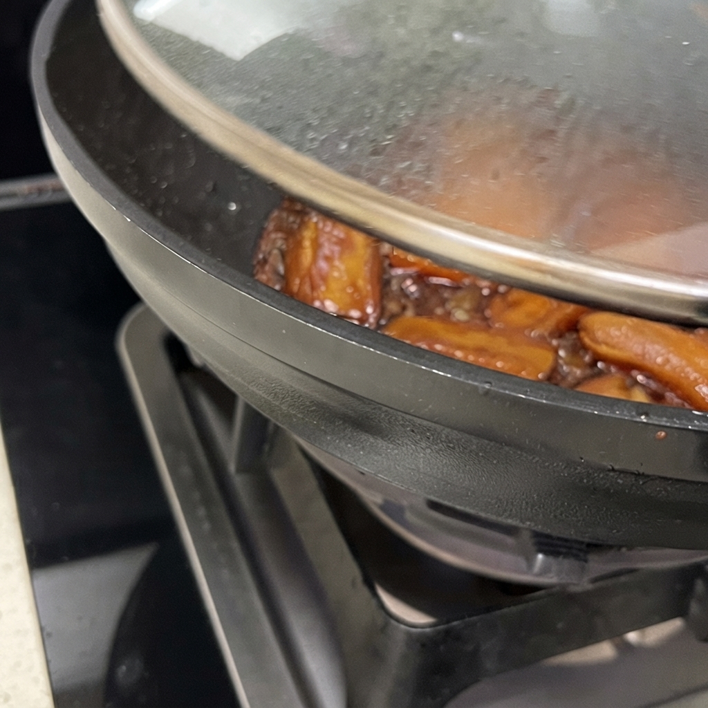 Nahaufnahme eines Woks mit halb geöffnetem Deckel, in dem Schweinerippchen in einer glänzenden, braunen Soße köcheln.