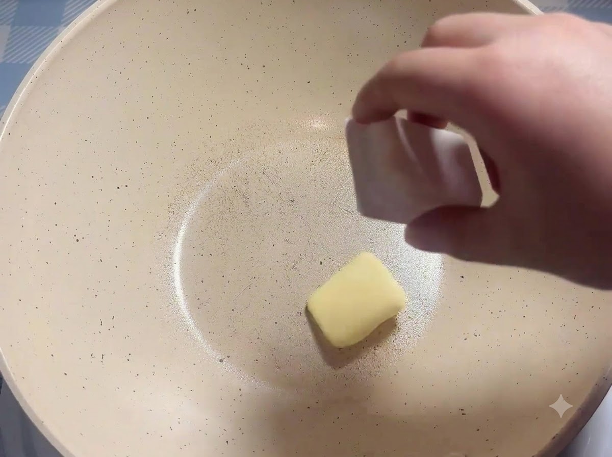 Ein Butterstück wird in eine erhitzte weiße Bratpfanne gegeben.