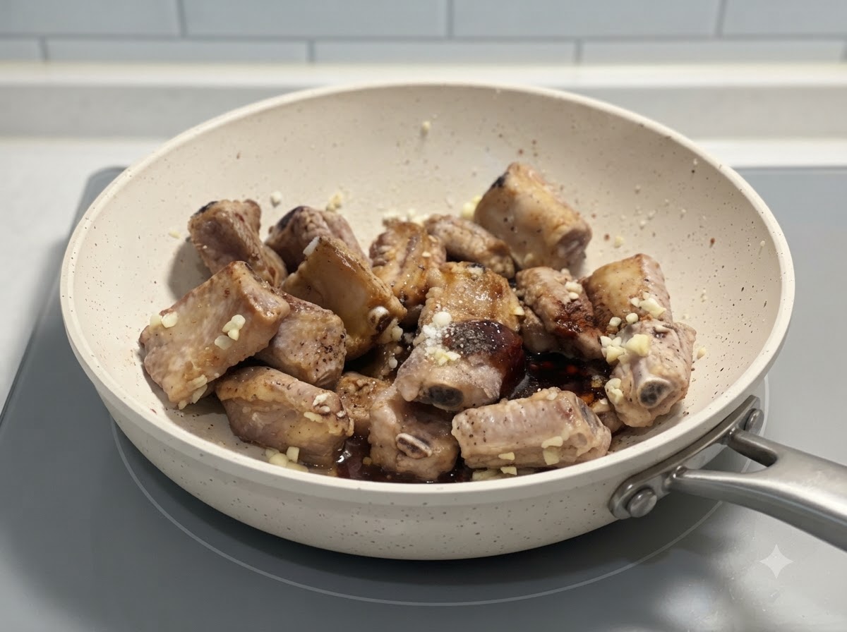 Schweinerippchen werden mit Gewürzen und gehacktem Knoblauch in einer weißen Pfanne auf einem Herd zusammengeschwenkt.