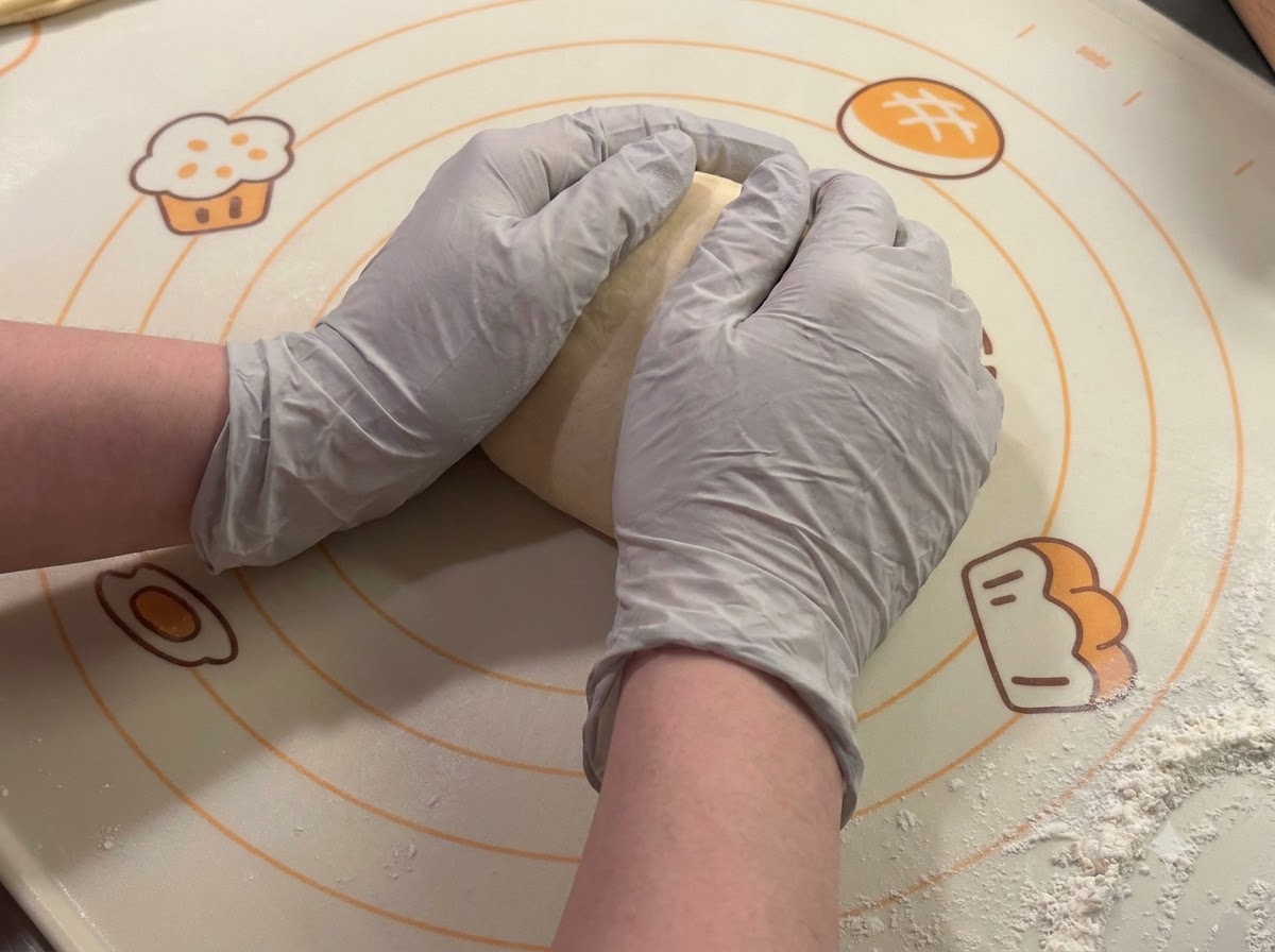 Behandschuhte Hände formen und runden Teigportionen auf einer Backmatte.