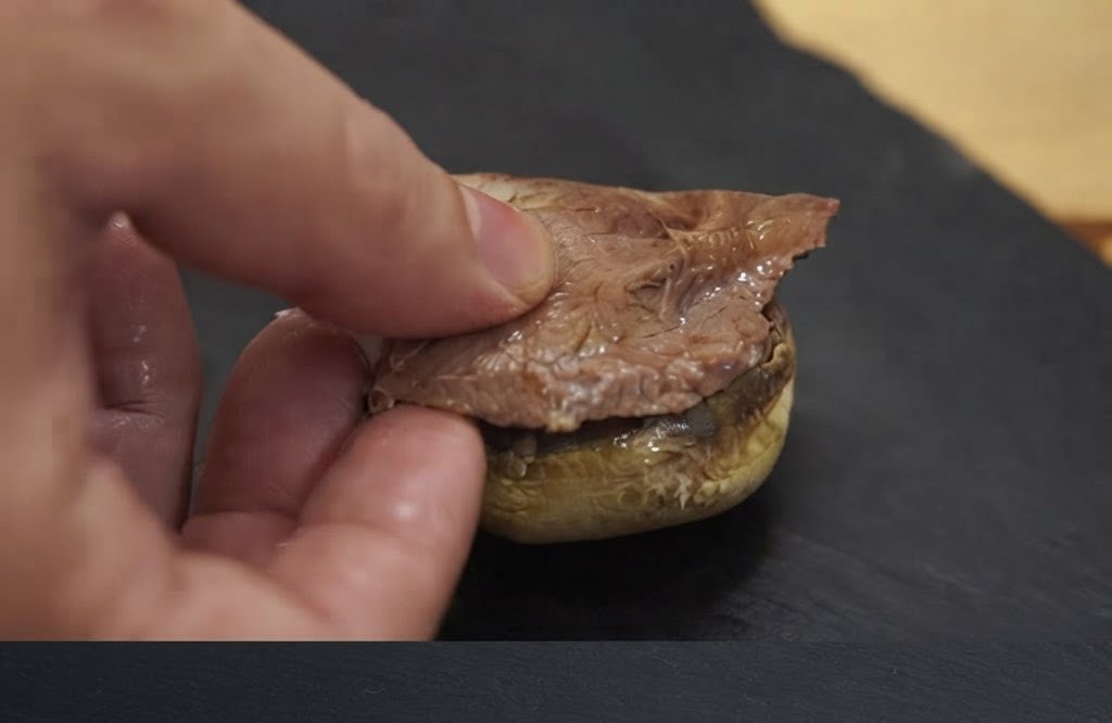 Eine Hand legt ein Stück gekochtes Fleisch auf eine gebackene Pilzkopfgrundlage.
