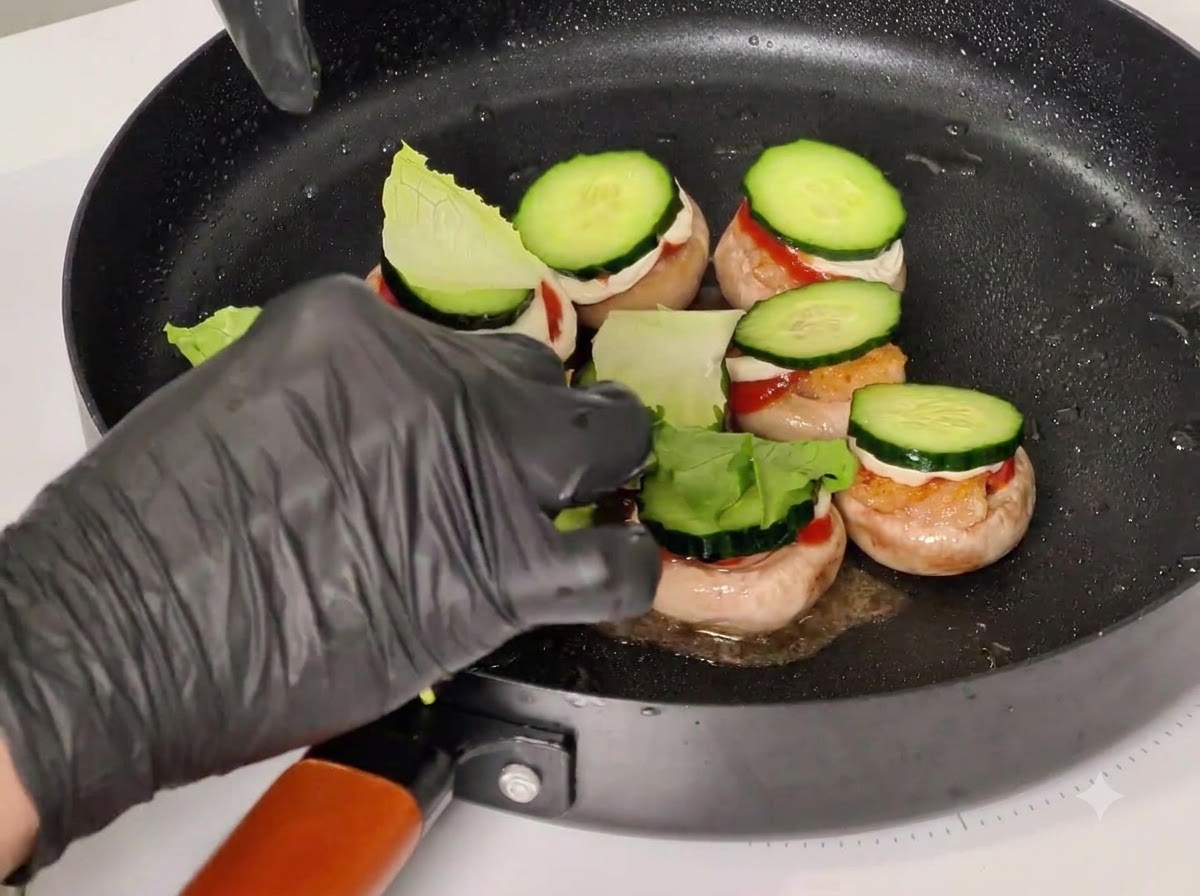 Eine handschuhbedeckte Hand steckt Zahnstocher durch gestapelte Zutaten in Pilzköpfen in einer schwarzen Bratpfanne.