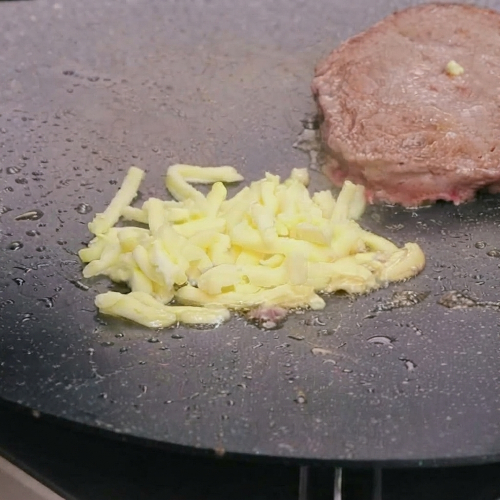 Geriebener Mozzarella-Käse schmilzt auf einem angebratenen Rinderpatty in einer heißen Pfanne.
