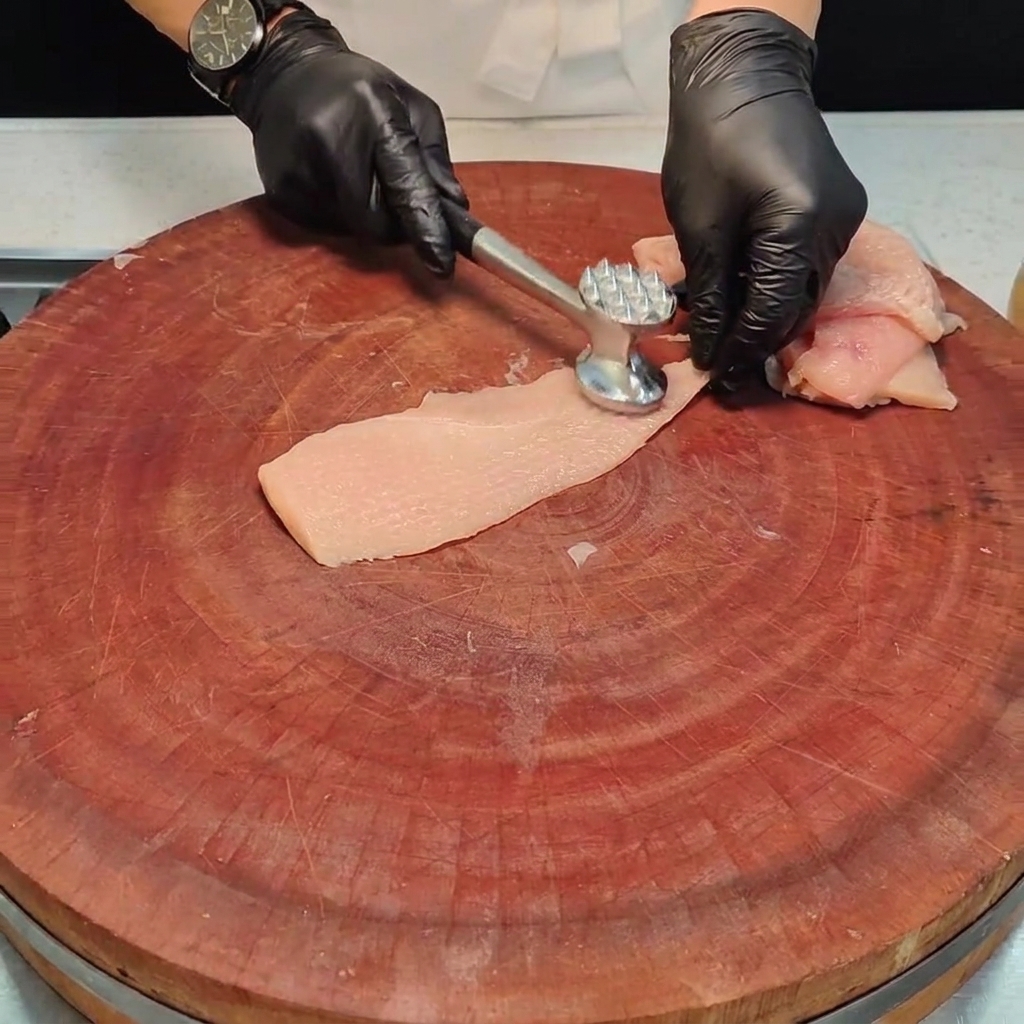 Ein Stück Hähnchenbrust wird mit einem Fleischklopfer aus Metall auf einem Holzbrett flach geklopft.