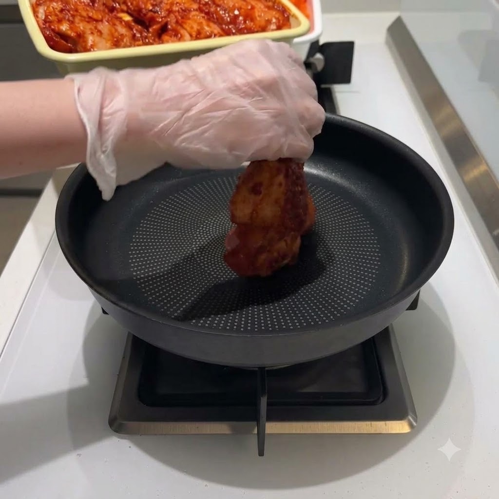 Eine behandschuhte Hand legt ein Stück rot mariniertes Hähnchen in die Mitte einer schwarzen Bratpfanne.