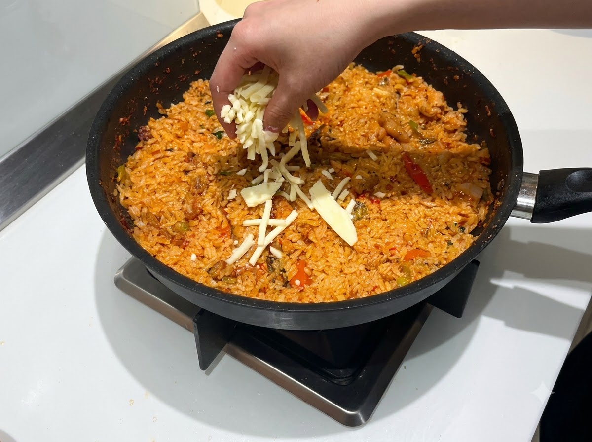 Eine Hand streut eine großzügige Menge geriebenen Mozzarella-Käses über eine Pfanne mit rotem, scharfem Bratreis.