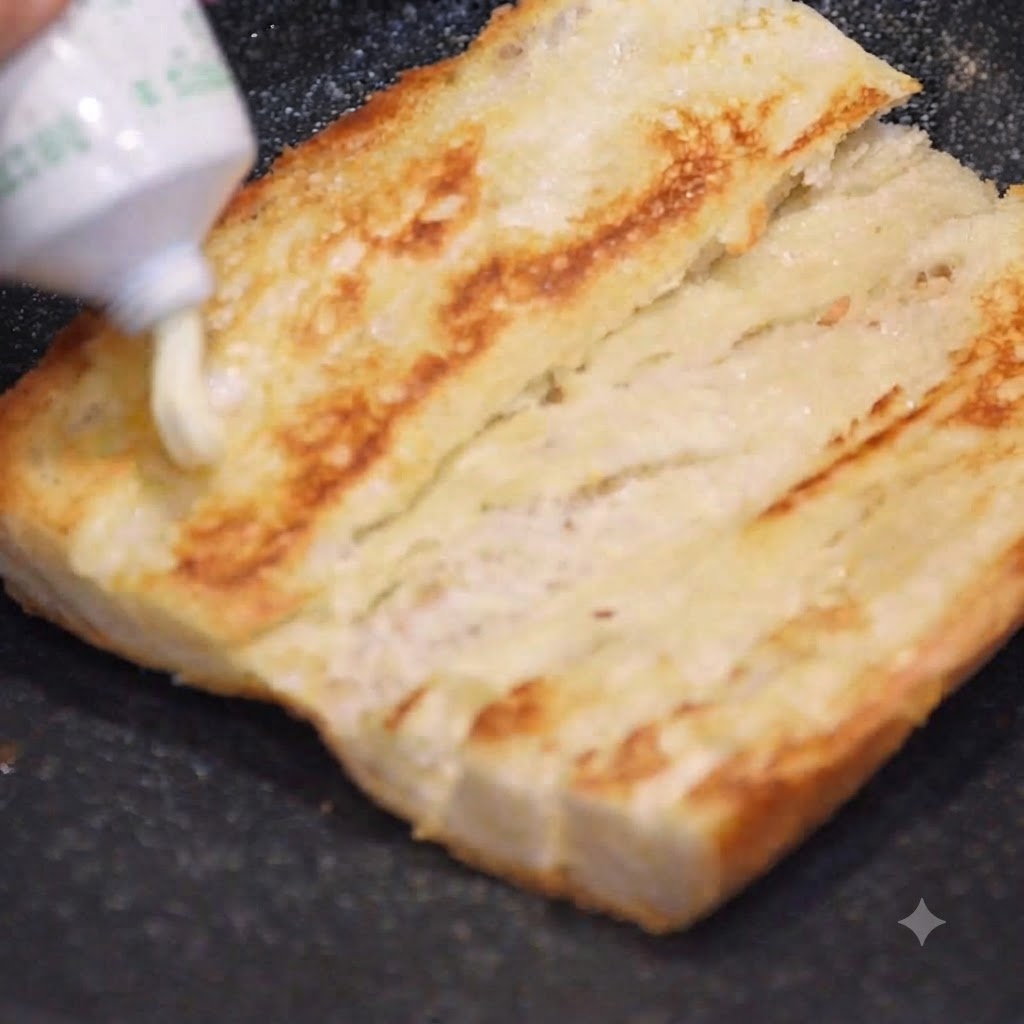 Mayonnaise wird aus einer Tube auf eine goldbraune Baguettehälfte gedrückt.