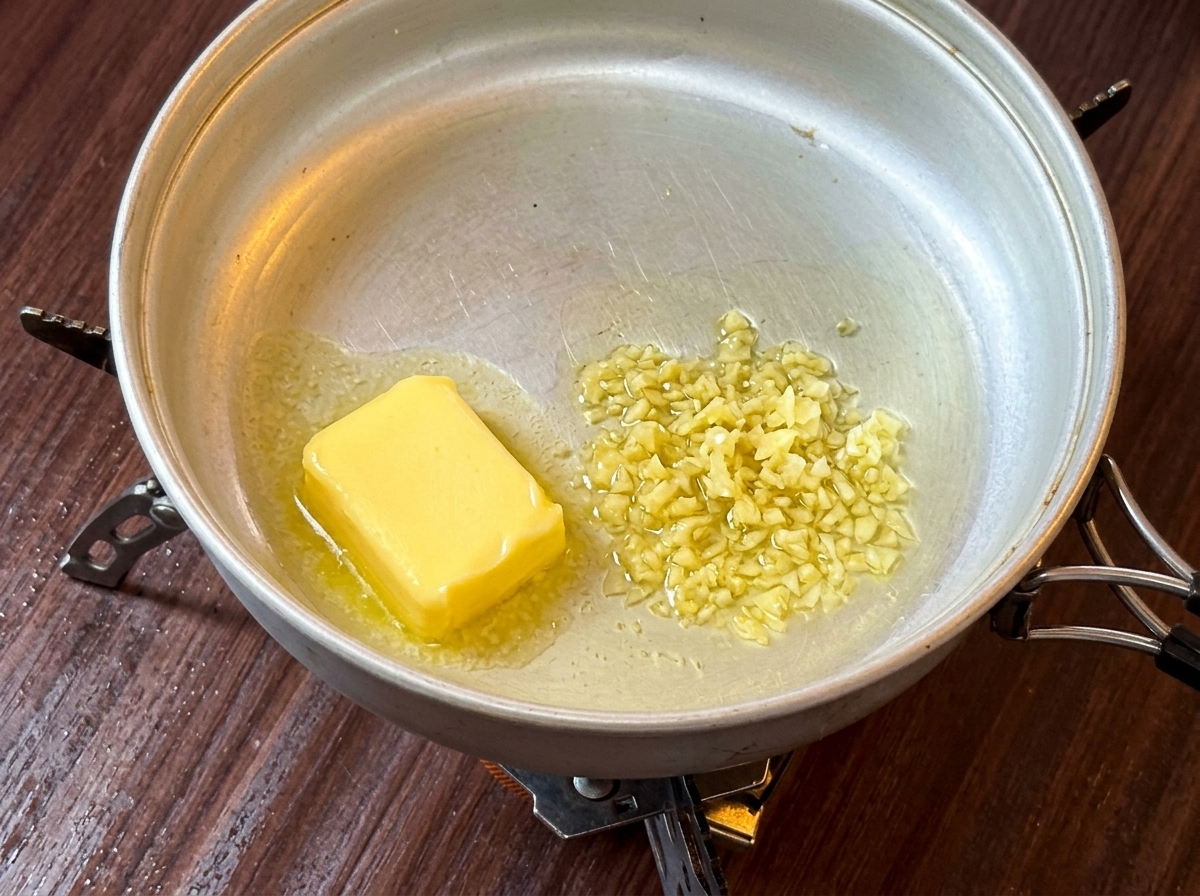 Ein Stück gelbe Butter schmilzt in einer Metallpfanne auf einem Campingkocher.