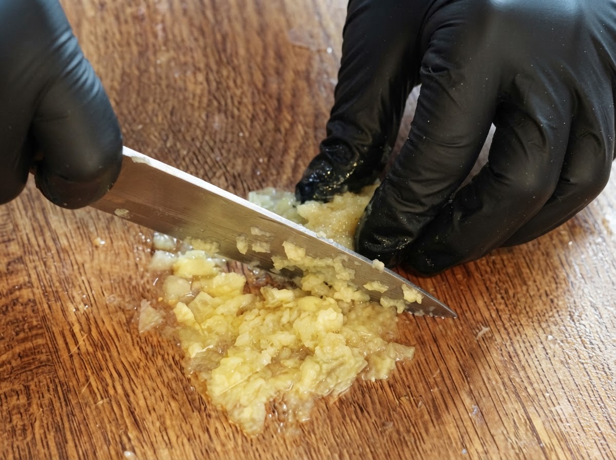 Ein Paar handschuhbedeckte Hände schneidet und püriert übrig gebliebenes Zitronenfleisch auf einem Holzschneidebrett mit einem Messer.